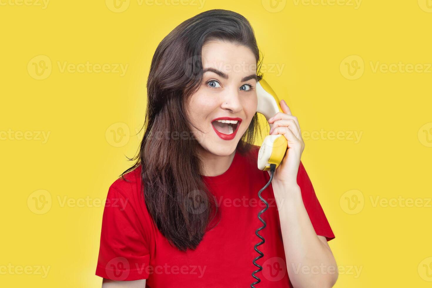 A beautiful brunette woman with red lipstick speaks on an old yellow telephone with a cord. She is smiling. on a yellow background photo