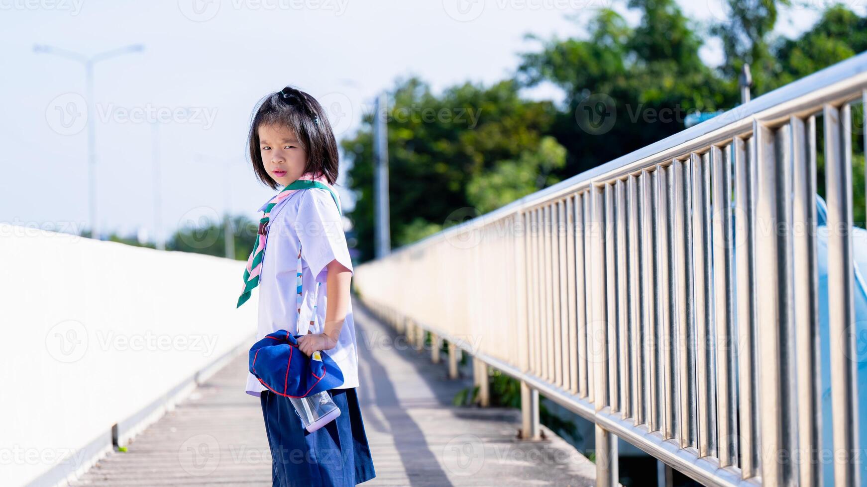 un Chica de escuela camina hogar desde colegio en el caliente verano sol, un niña frunce el ceño en contra el caliente sol, niño camina en un paso superior, mira espalda a el cámara, un 6 6 año antiguo niño usa un tailandés colegio uniforme. foto