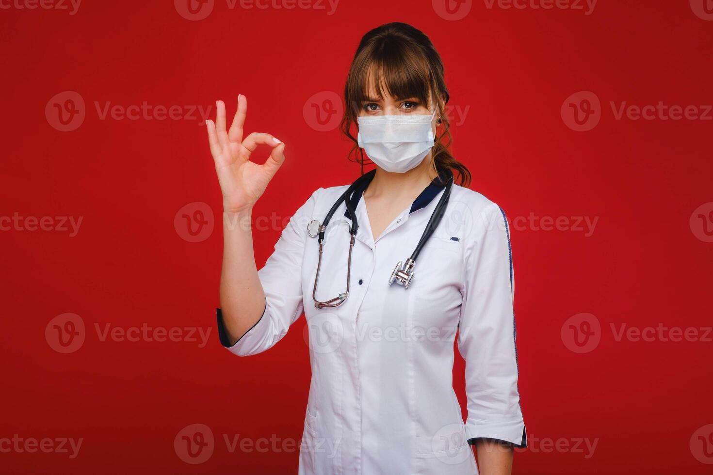 A young doctor in a white coat and medical mask stands on a red background photo