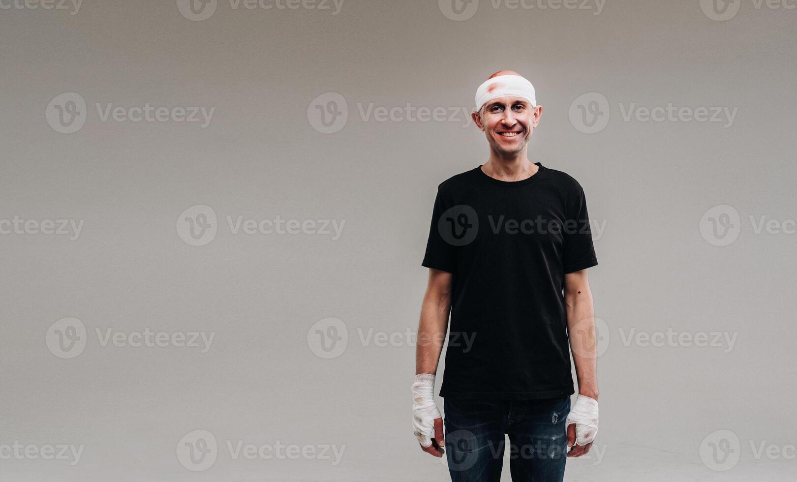 Against a gray background stands a battered and battered man in a black T-shirt with his head wrapped in a bandage photo