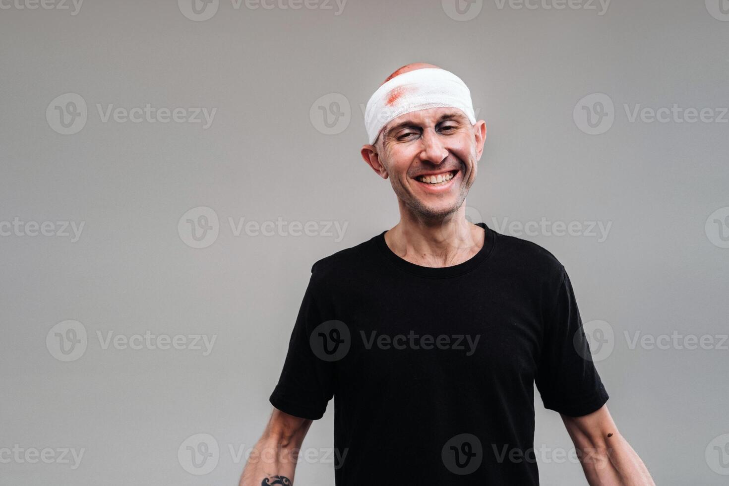 Against a gray background stands a battered and battered man in a black T-shirt with his head wrapped in a bandage photo