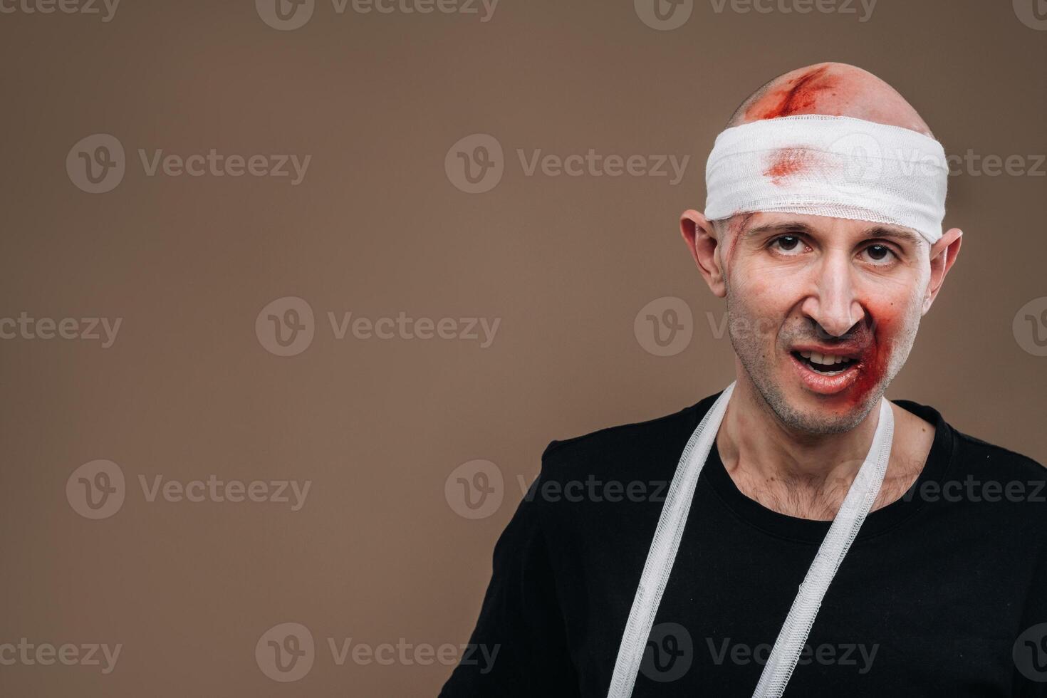 A battered man with a bandaged head and a cast on his arm stands on a gray background photo