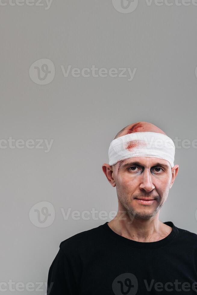 Against a gray background stands a battered and battered man in a black T-shirt with his head wrapped in a bandage photo