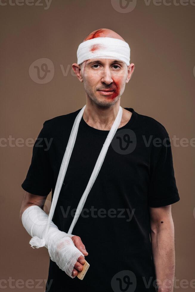 A battered man with a bandaged head and a cast on his arm stands on crutches on a gray background photo