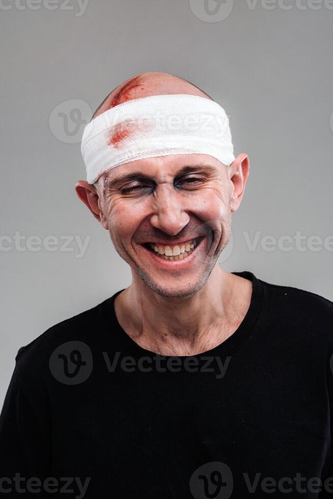 Against a gray background stands a battered and battered man in a black T-shirt with his head wrapped in a bandage photo