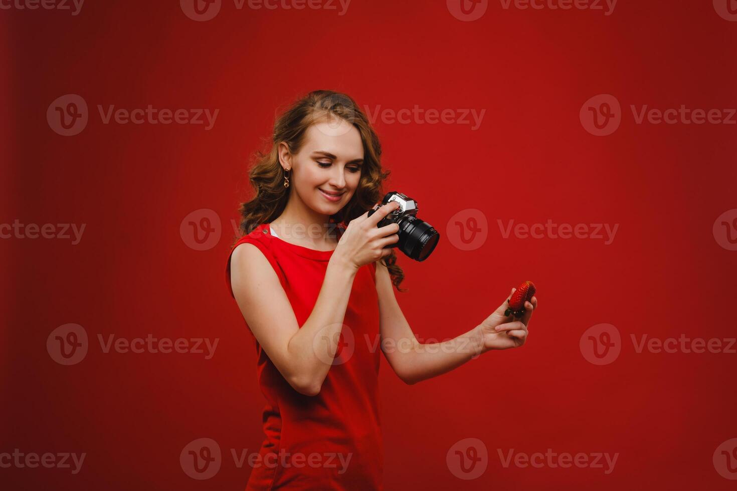 a smiling young woman with wavy hair holds a strawberry and photographs it, holding a delicious fresh strawberry on a bright red background photo
