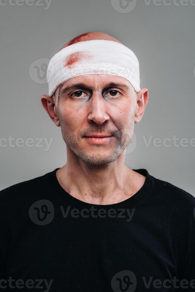 Against a gray background stands a battered and battered man in a black T-shirt with his head wrapped in a bandage photo