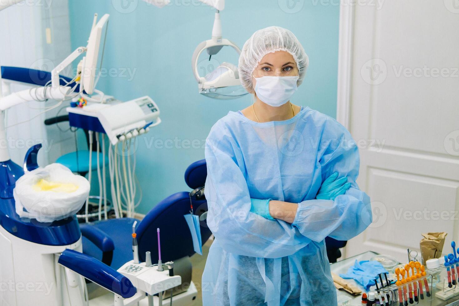 un hembra dentista vistiendo un médico máscara y caucho guantes poses para el cámara y pliegues su brazos en su oficina foto