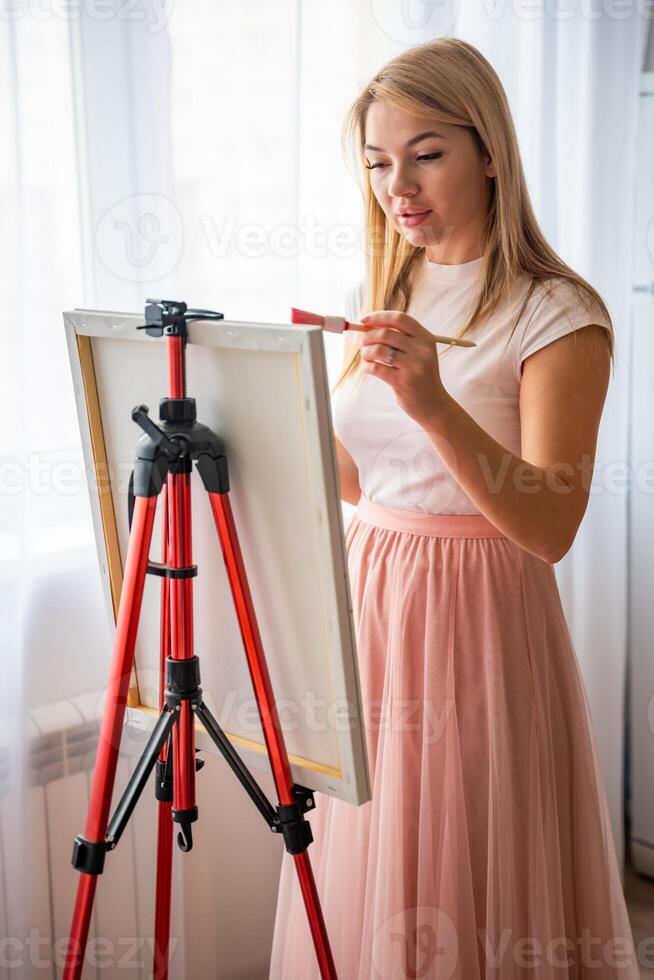 Beautiful young woman artist with palette and brush painting abstract pink picture on canvas. Art and creativity concept photo