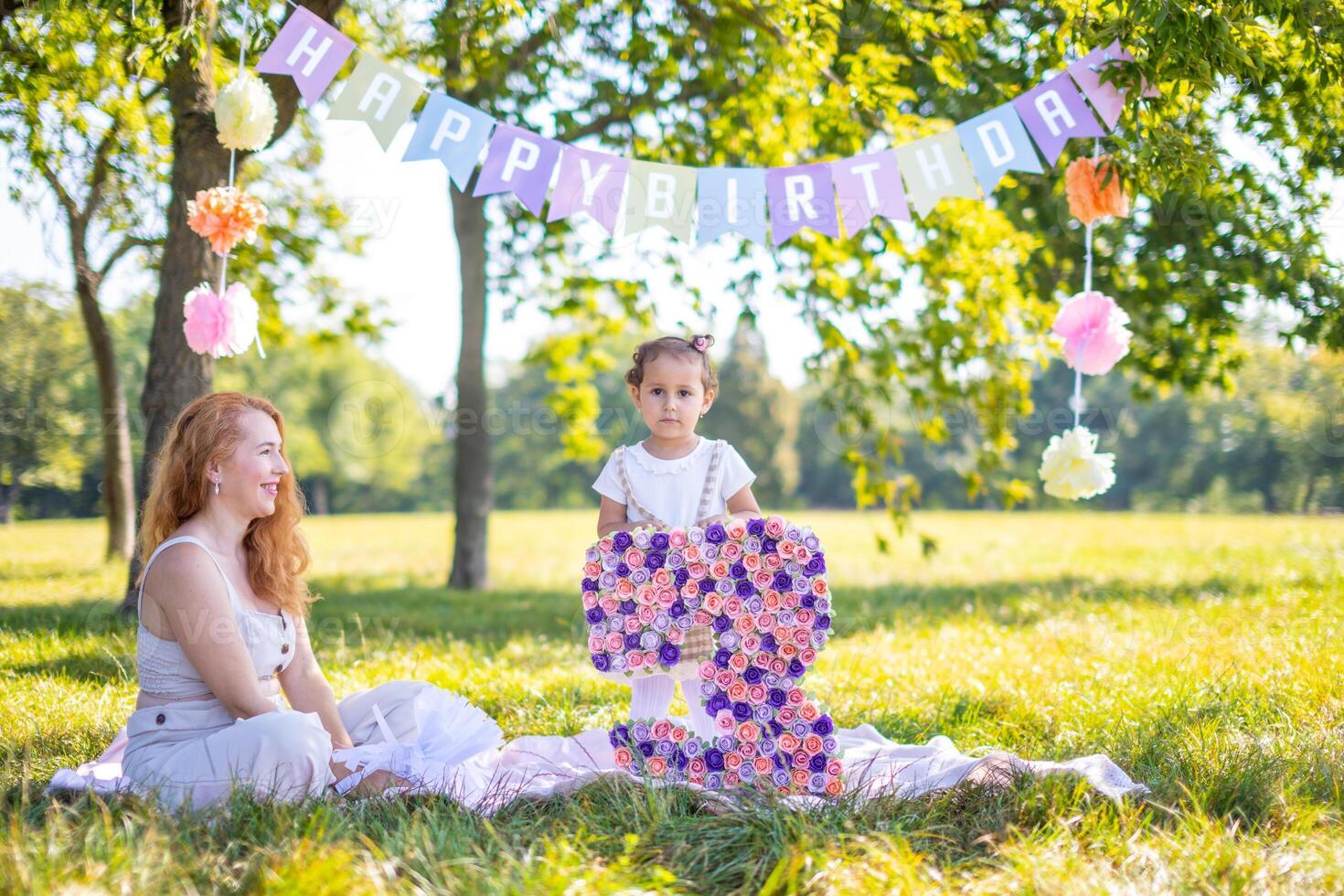 Cheerful mother and daughter having fun on child birthday on blanket with paper decorations in the park photo