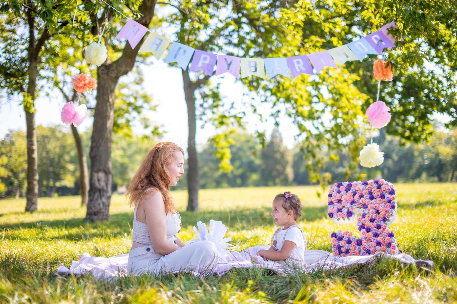 Cheerful mother and daughter having fun on child birthday on blanket with paper decorations in the park photo