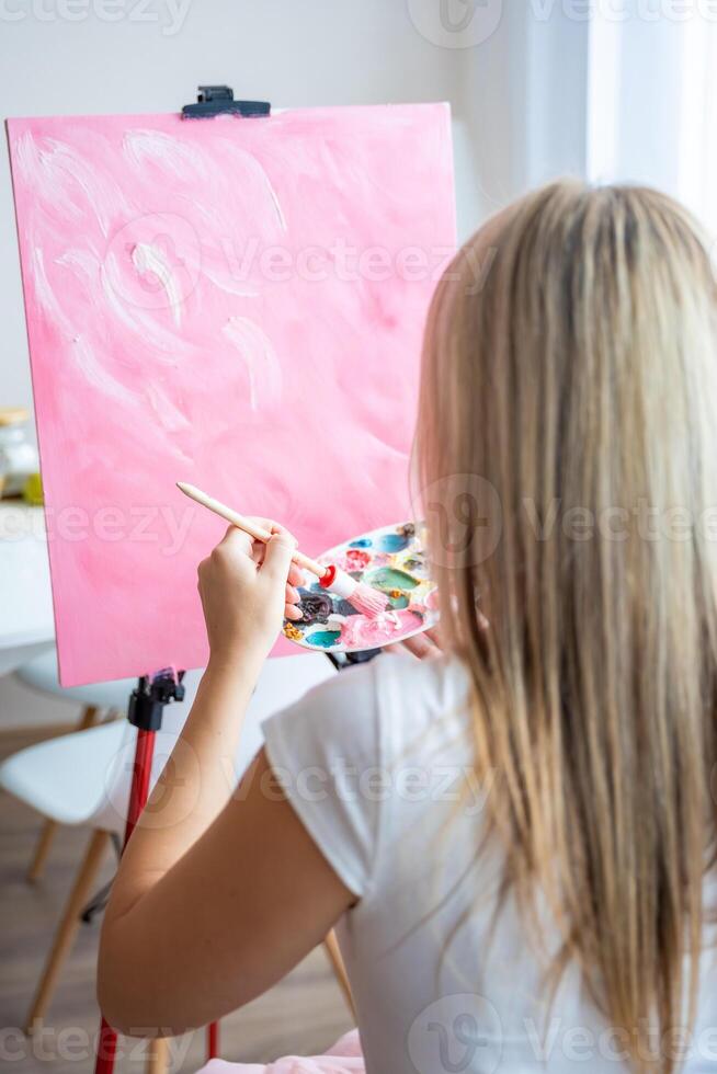 espalda ver de joven mujer artista con paleta y cepillo sentado y pintura resumen rosado imagen en lona cerca ventana a hogar. Arte y creatividad concepto foto