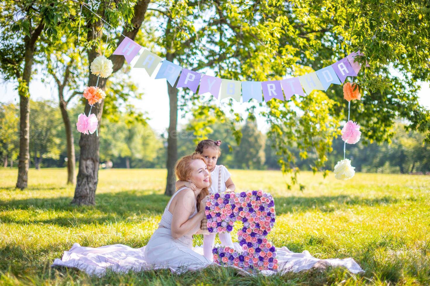 Cheerful mother and daughter having fun on child birthday on blanket with paper decorations in the park photo