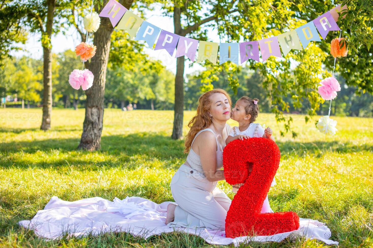 Cheerful mother and daughter having fun on child birthday on blanket with paper decorations in the park photo