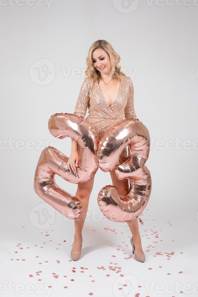 Pretty sexy blonde woman on white background. Portrait of beautiful young lady in beige glitter sequins long evening dress with twenty five shape inflatable balloons in hand. photo