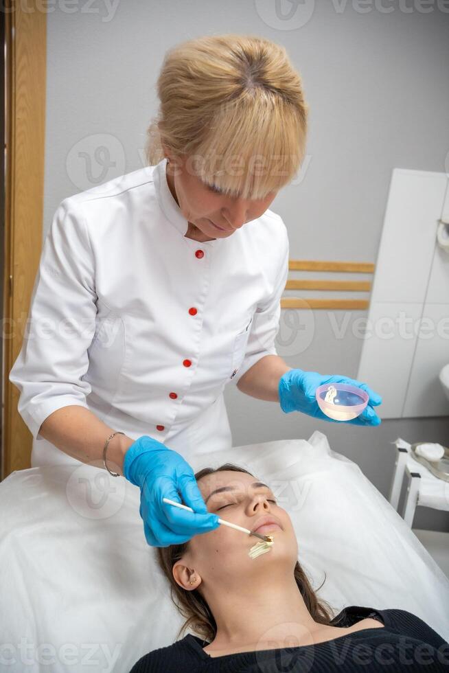 Doctor cosmetologist or dermatologist making face mask in cosmetology salon. Professional Beautician applying face mask on caucasian woman face lying on bed in bathrobe. photo