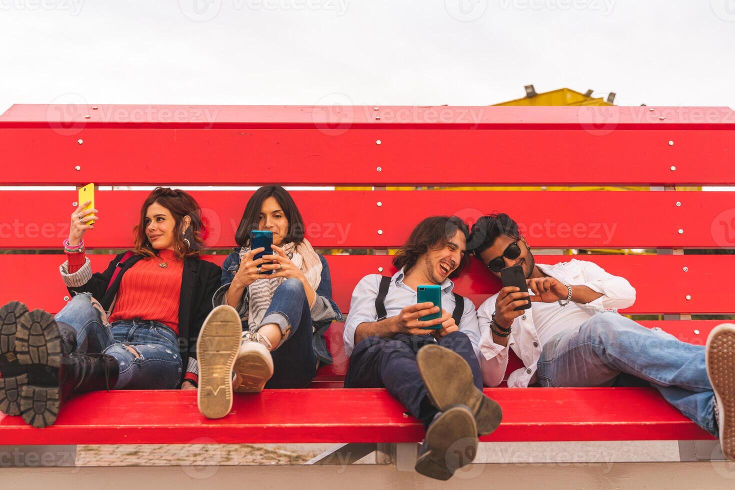 group of multi-cultural best friends sitting on a big red bench use smart phones photo