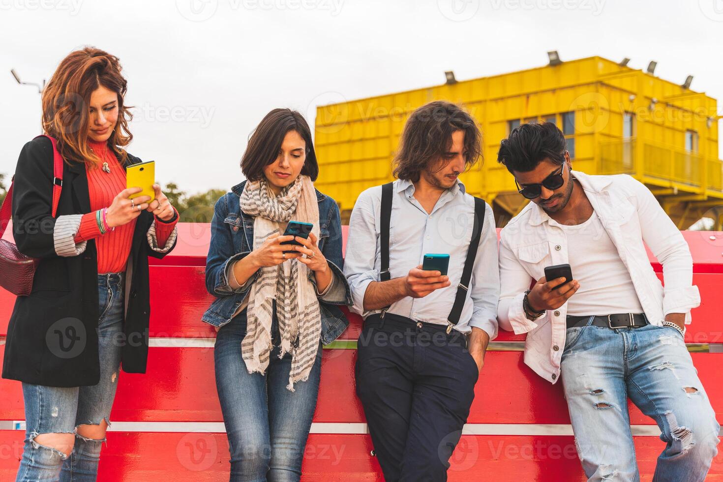 group of multi-cultural best friends sitting on a big red bench use smart phones photo