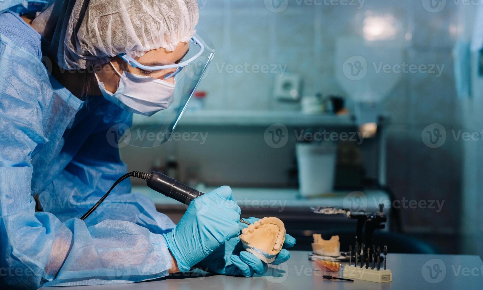 A masked and gloved dental technician works on a prosthetic tooth in his lab photo