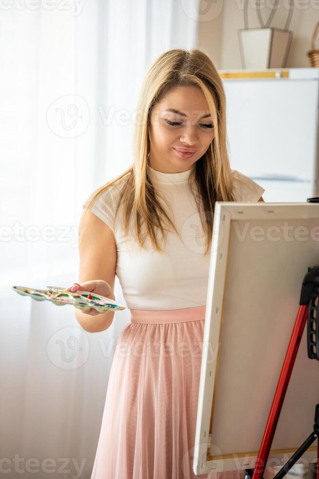 Young woman artist with palette and brush painting abstract pink picture on canvas at home. Art and creativity concept photo