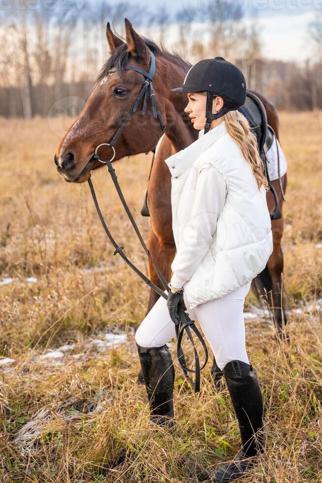 rubio profesional hembra jockey en pie cerca caballo en campo en invierno. amistad con caballo foto