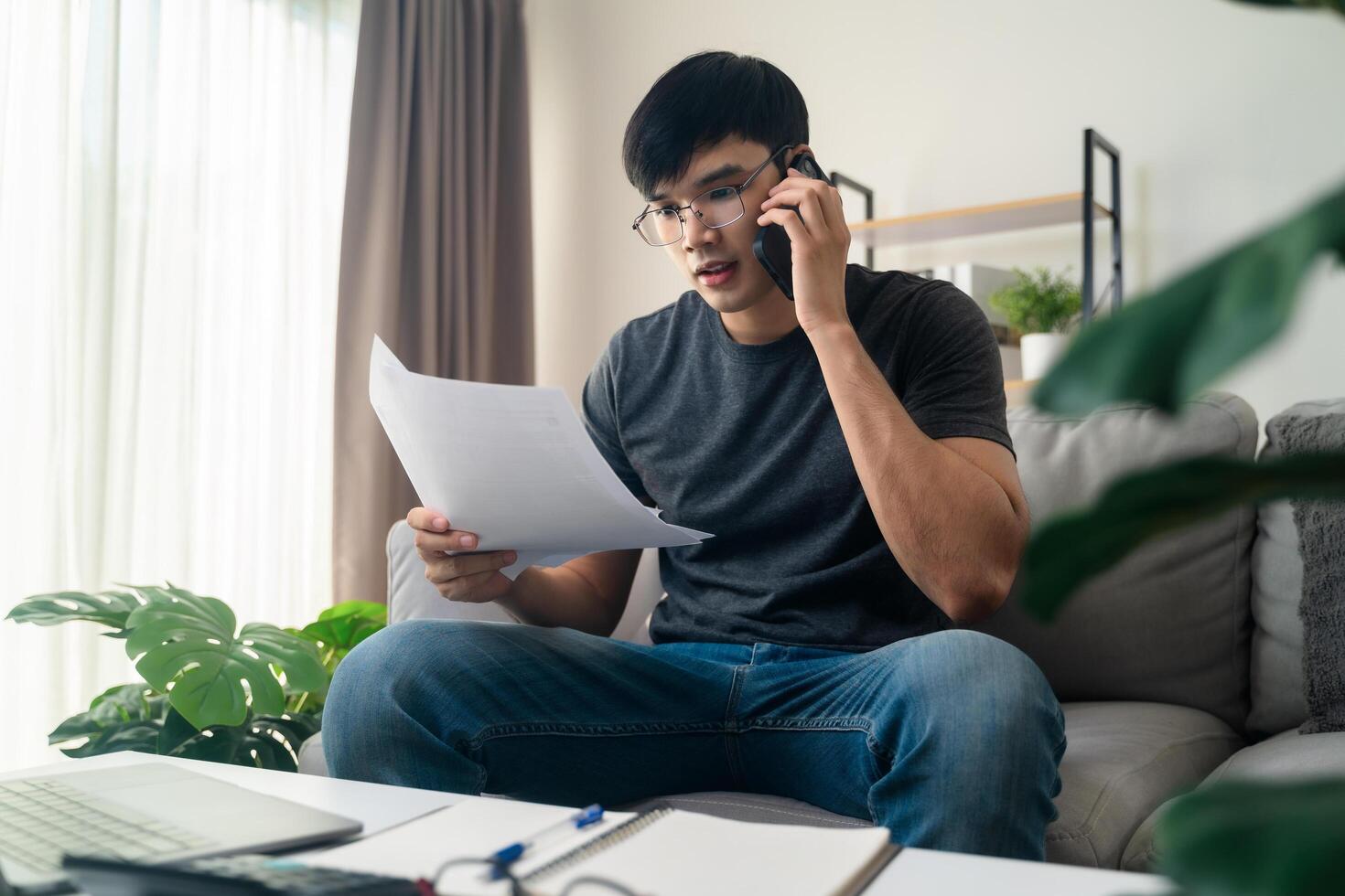 the man in casual clothes working with a laptop, computer, smart phone, calculator sitting on the sofa in the living room at home, working from home concept. photo