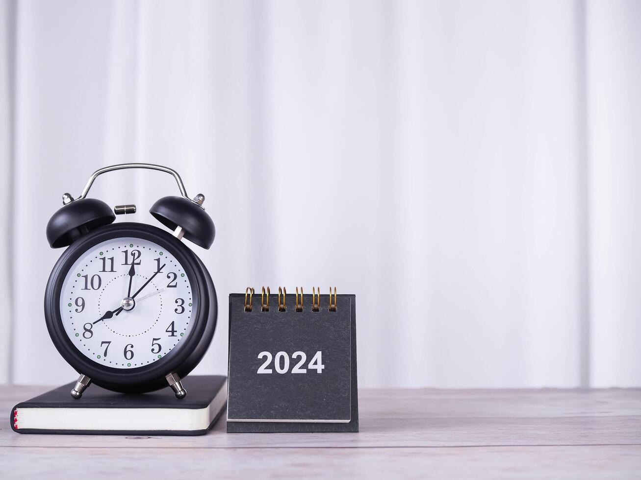 Creative desk with 2024 calendar, Black alarm clock on hardcover book with white curtain for background. Copy space for text, Office desk concept. photo