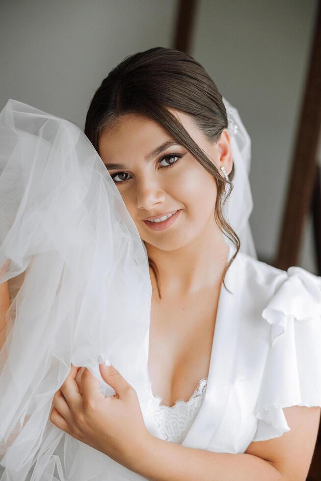 portrait of an incredibly beautiful girl bride in a white robe in the bedroom, the bride poses holding her veil in her hands and covers herself with it. photo