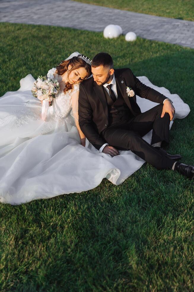 Portrait of beautiful wedding couple sitting on green grass in park in summer. Young cheerful woman bride hugging groom. photo