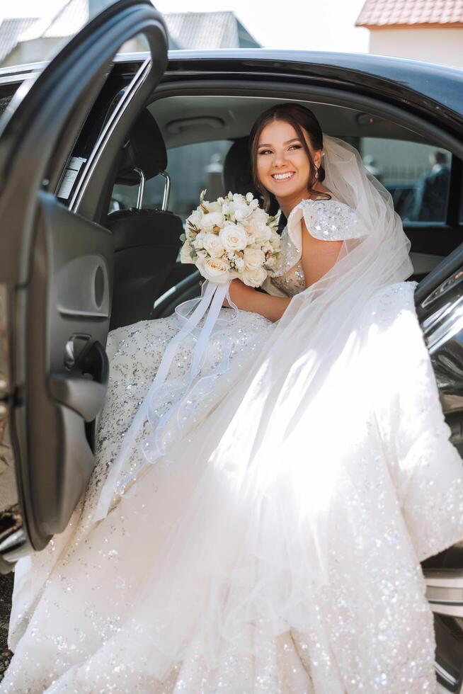 el novia es sentado en un negro coche con un ramo de flores de flores en su manos y mira fuera de el coche. retrato de un bastante tímido novia en un coche. novia, sonrisa, emociones foto