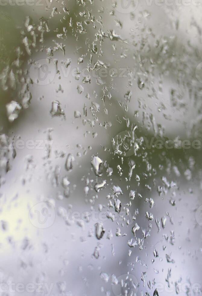 A photo of rain drops on the window glass with a blurred view of the blossoming green trees. Abstract image showing cloudy and rainy weather conditions