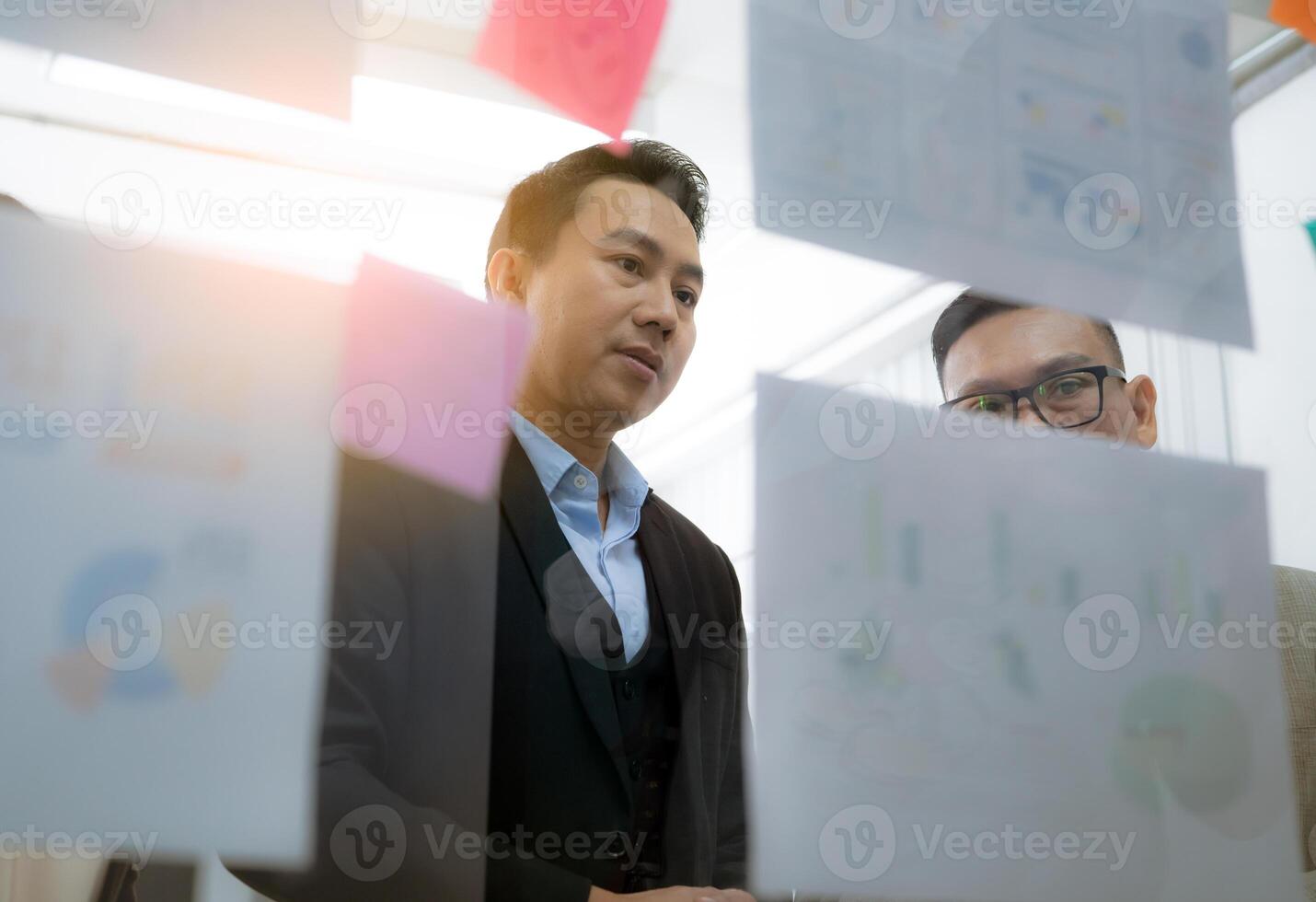 Two men are looking at a wall covered in paper with graphs and charts. They are discussing the information on the wall photo