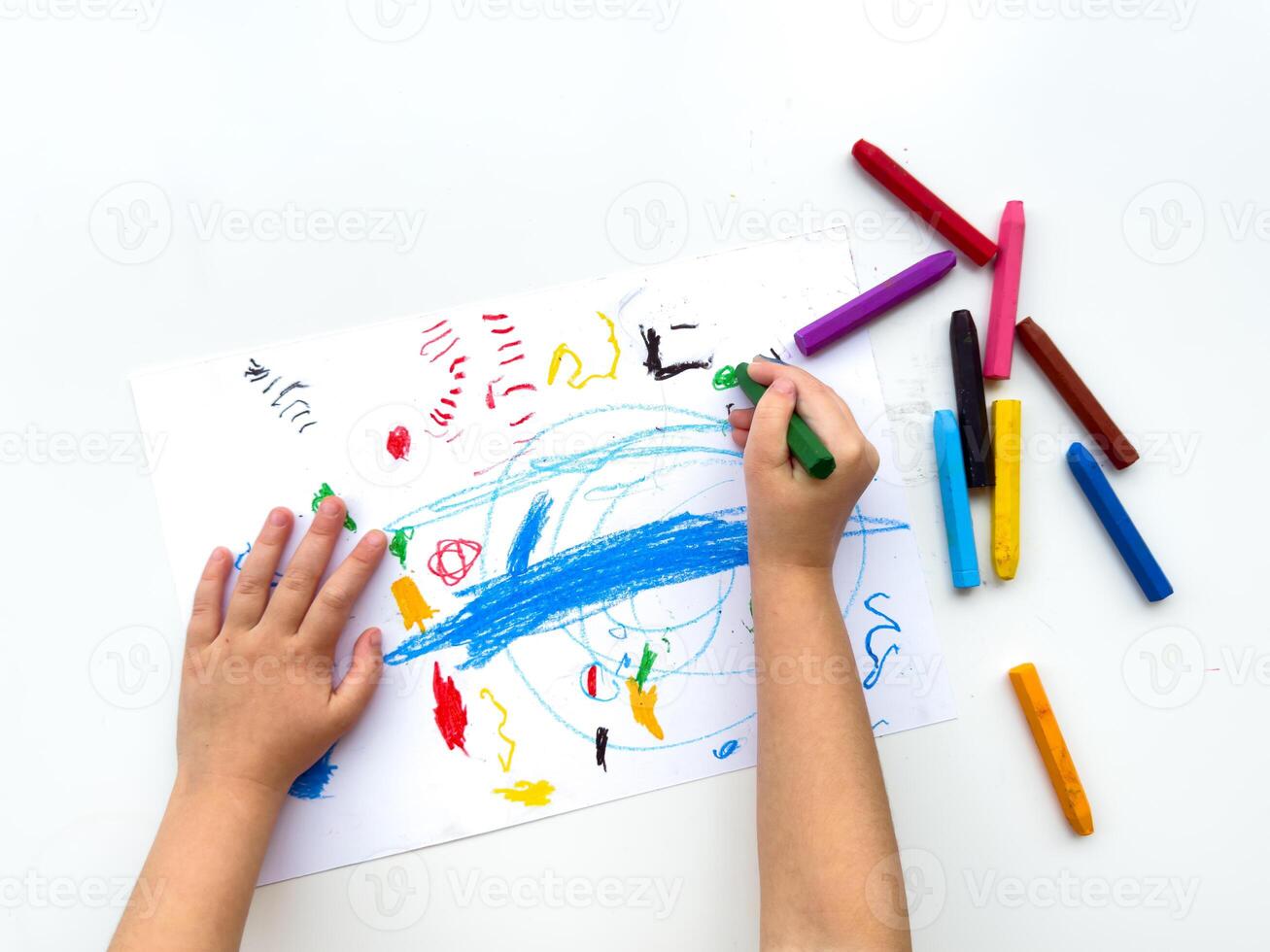 Childs hands drawing with colorful wax crayons on white paper, top view. Creative art concept for educational and developmental activities. photo