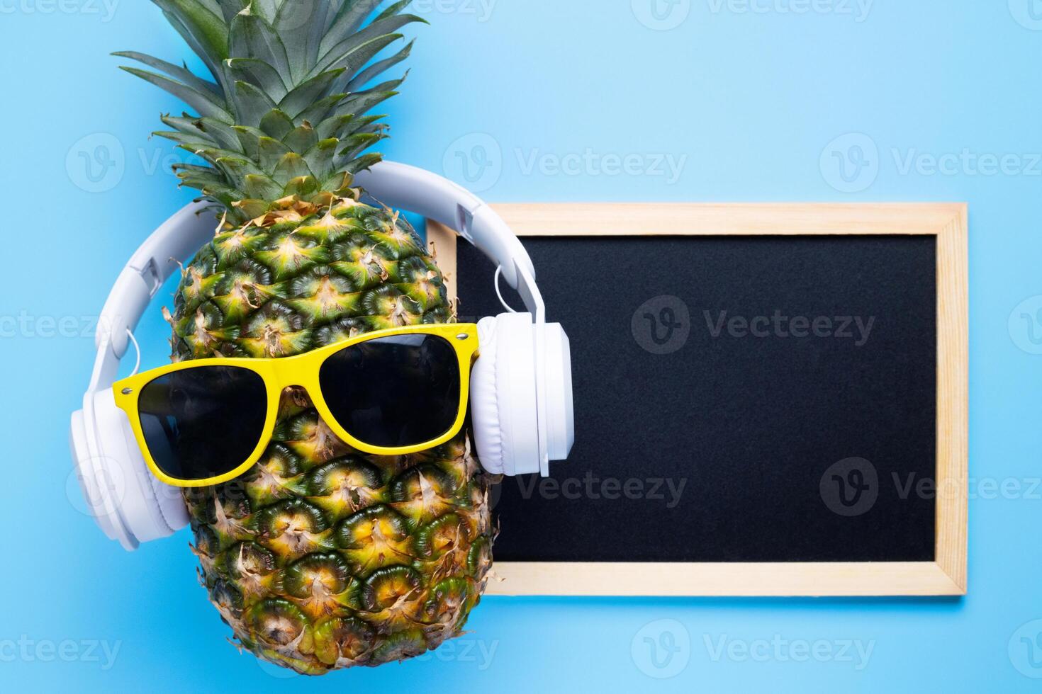 Pineapple in sunglasses and headphones with empty blackboard on blue background photo