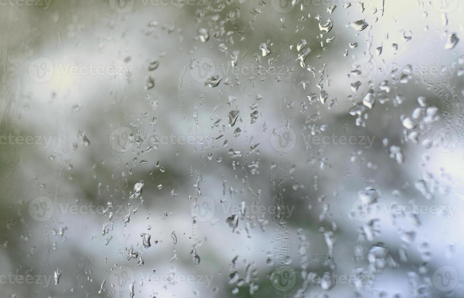 A photo of rain drops on the window glass with a blurred view of the blossoming green trees. Abstract image showing cloudy and rainy weather conditions