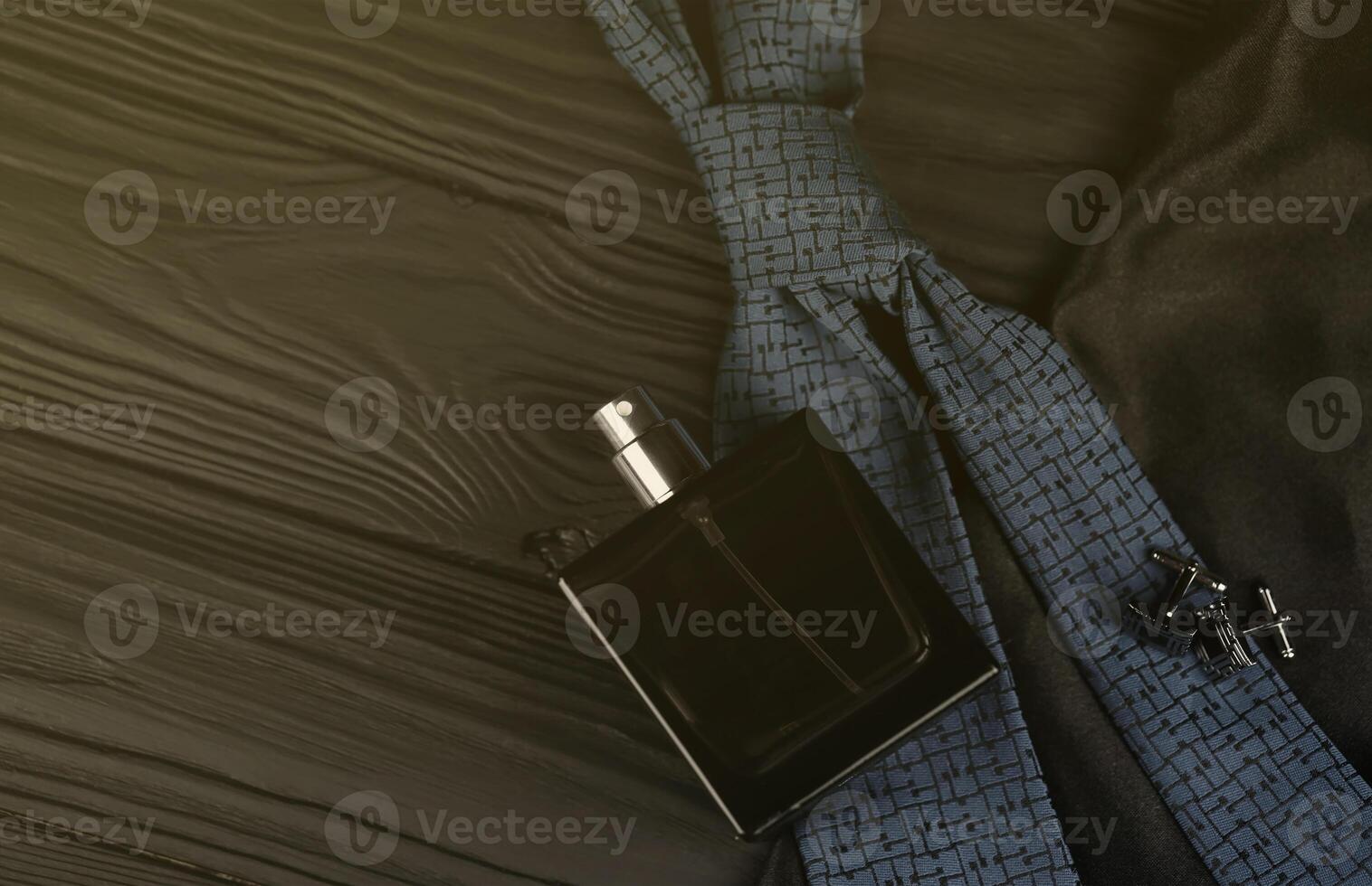 A bottle of mens cologne and cufflinks with blue tie lie on a black luxury fabric background on a wooden table. Mens accessories photo