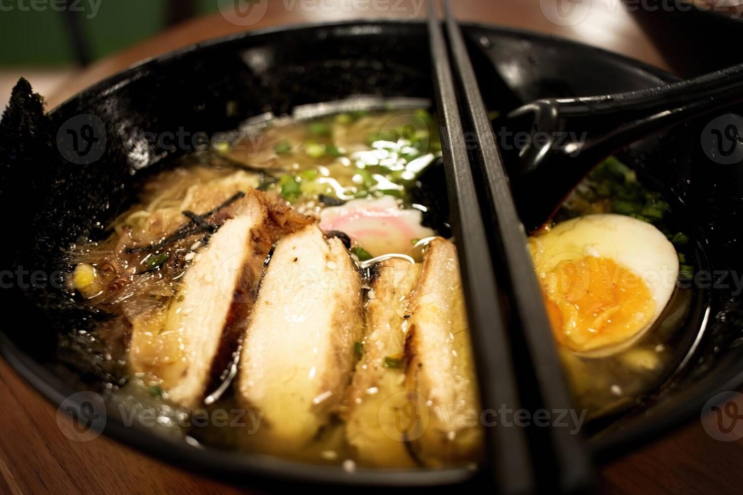 A bowl of ramen looks tempting with a golden yellow broth, topped with sliced tempura chicken, half-boiled egg, nori, mushrooms and finely chopped green onions. photo