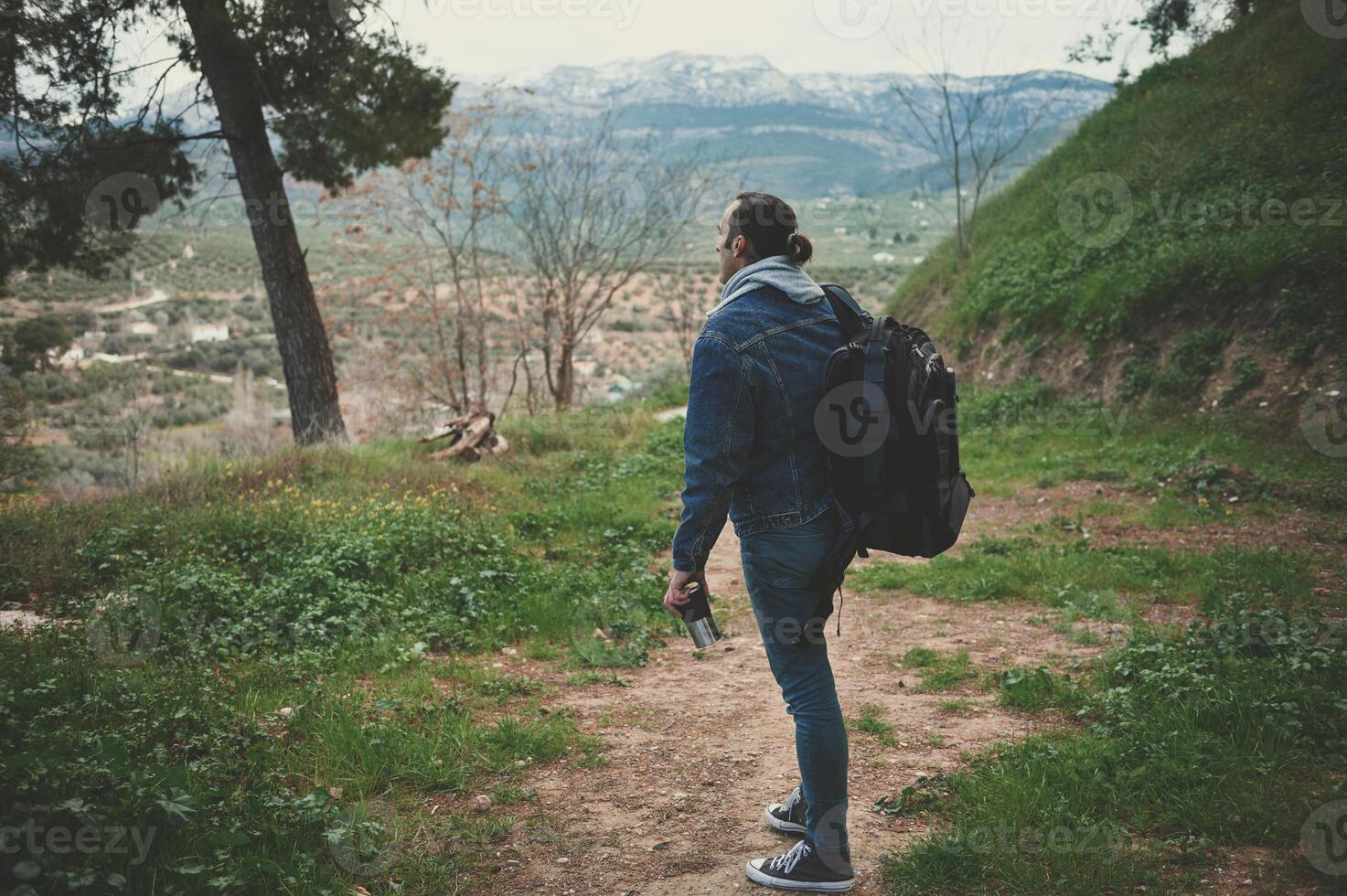 Full size dramatic shot of a hiker young man with backpack standing in the forest path and looking at the mountains photo