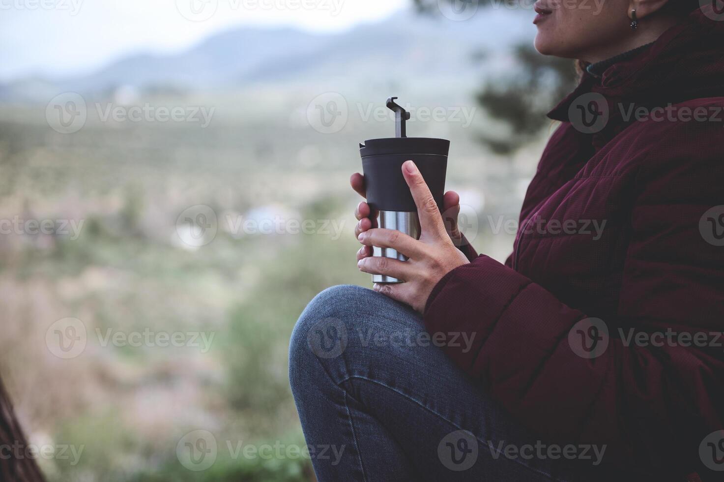 Female traveler holding thermos mug with hot drink, warming hands while relaxing after hiking in the forest or mountains photo