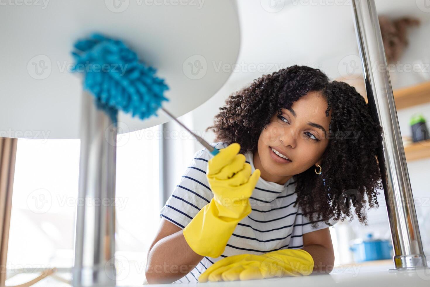 Young housekeeper in apron dusting the shelf by duster carefully cleaning in living room at home. young wife in rubber gloves doing housework. photo