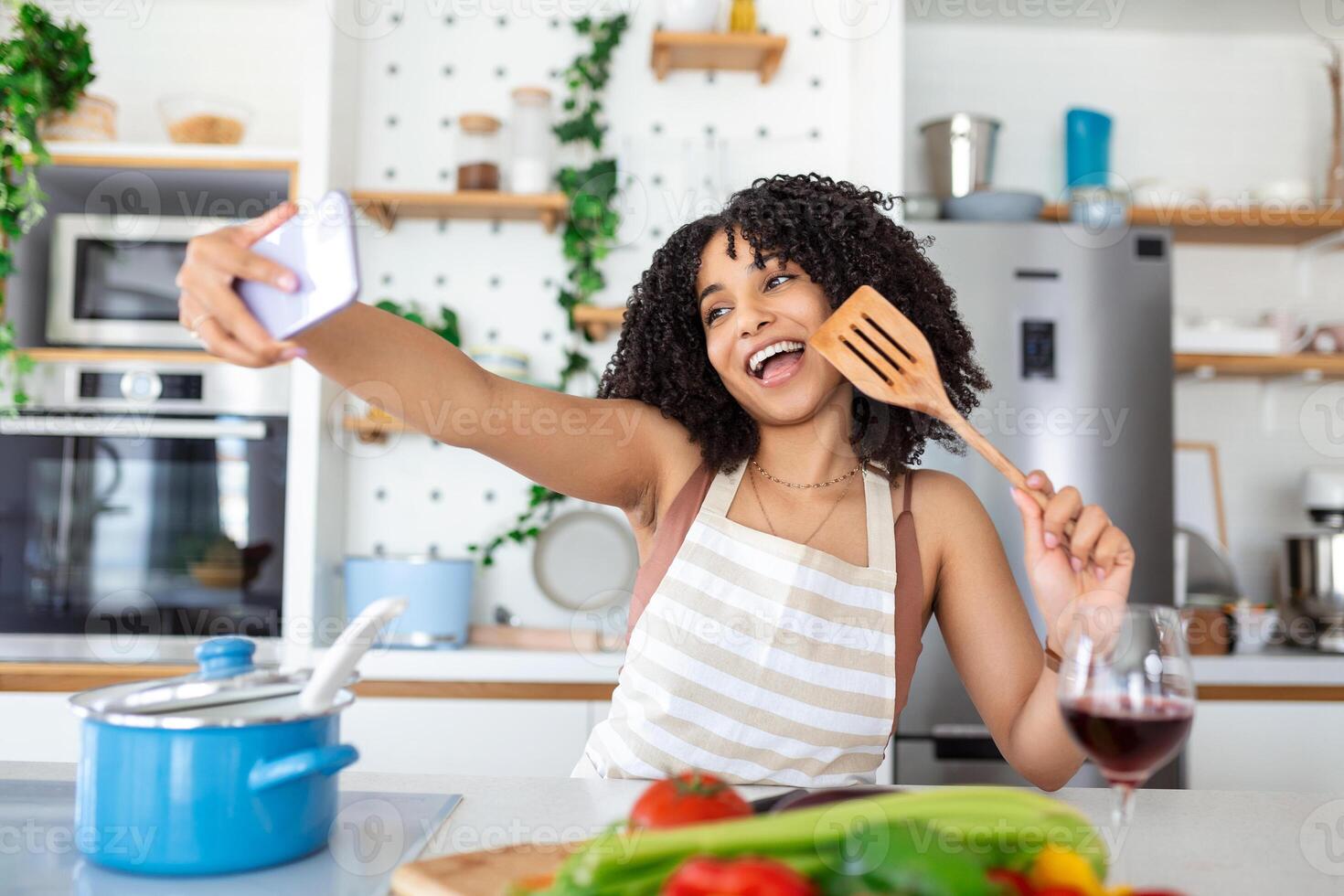 Portrait of smiling young African American woman taking selfie with smartphone while cooking in kitchen at home photo
