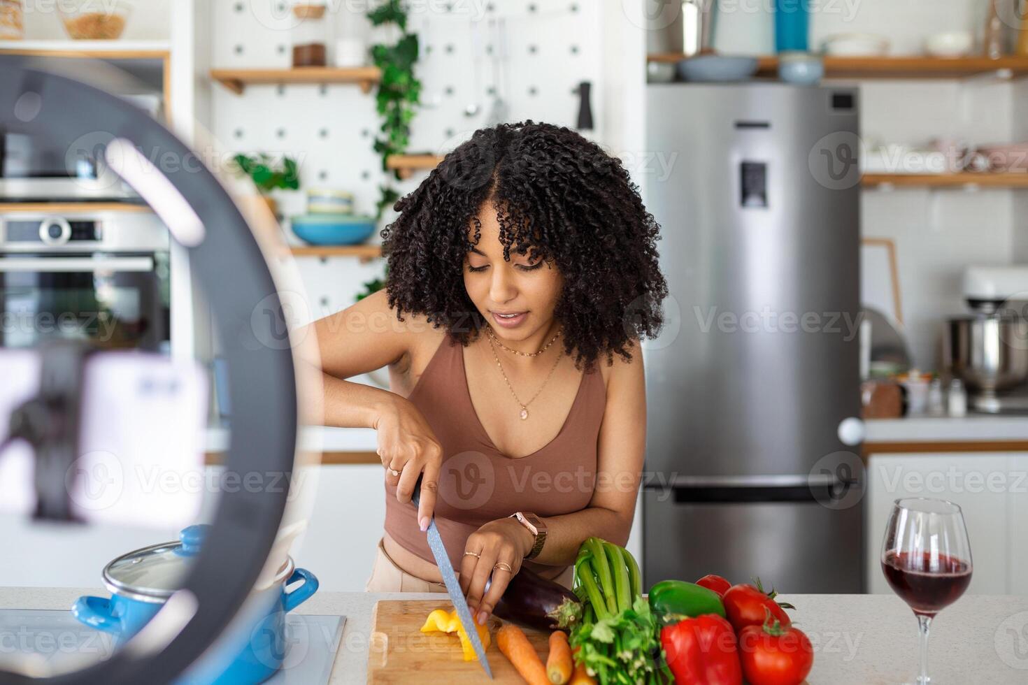 Cheerful food blogger wearing apron standing at table in modern kitchen cutting vegetables, shooting this process on camera photo