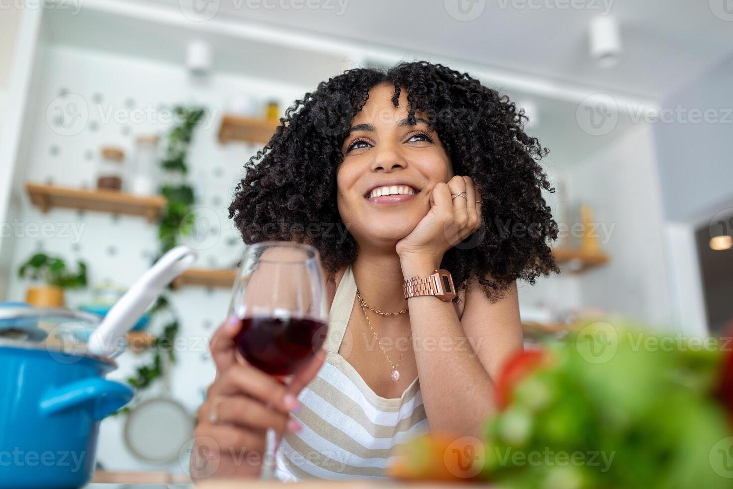 Smiling young housewife with red wine glass as she stands at the stove in the kitchen preparing dinner photo