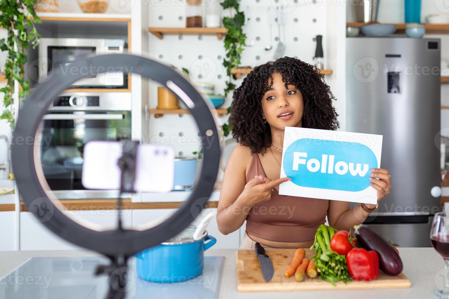 Happy african american woman vlogger broadcasting live video online while cooking food in kitchen at home holding follow sign photo