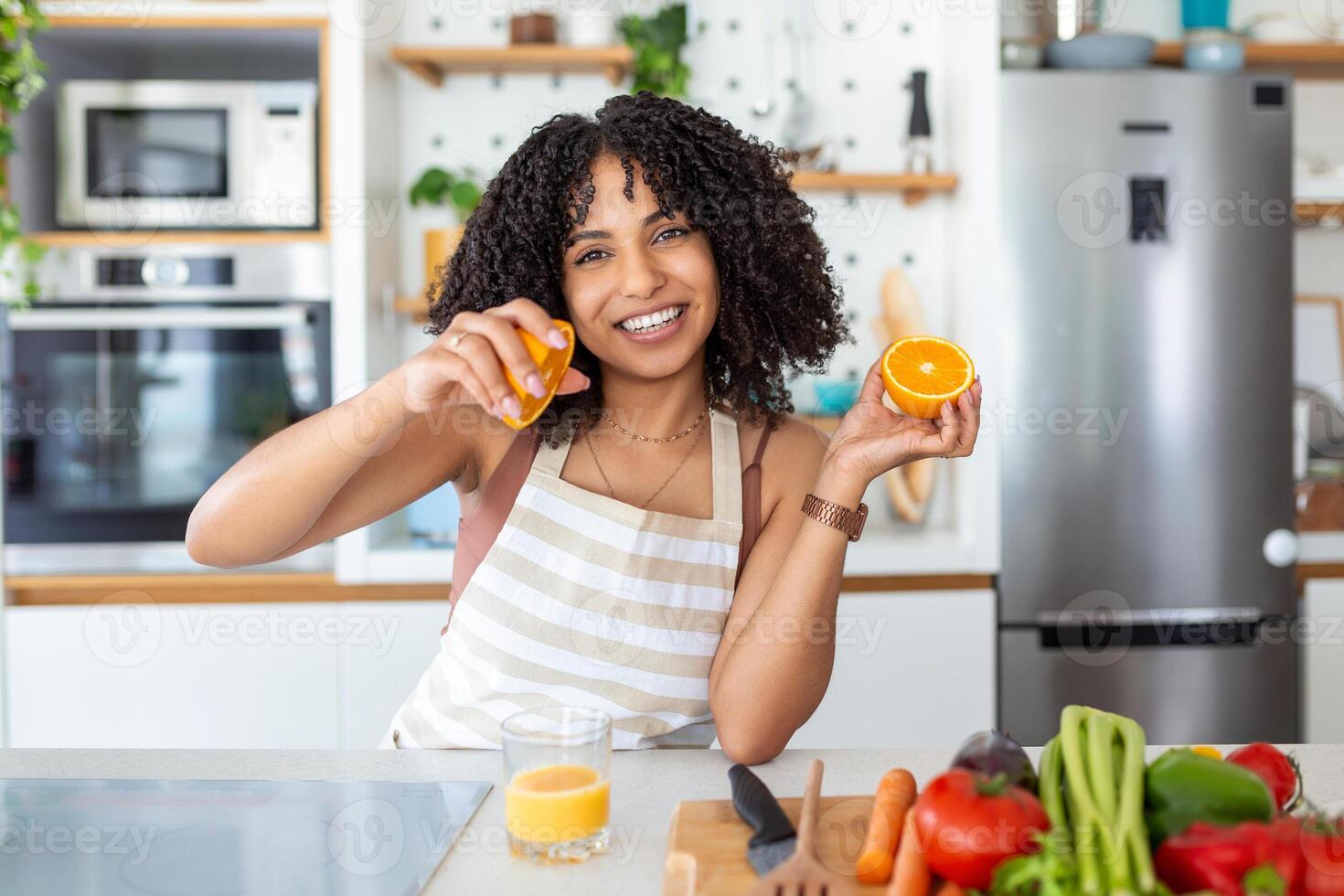 hermosa joven mujer Bebiendo Fresco naranja jugo en cocina. sano dieta. contento joven mujer con vaso de jugo y naranja a mesa en cocina. foto