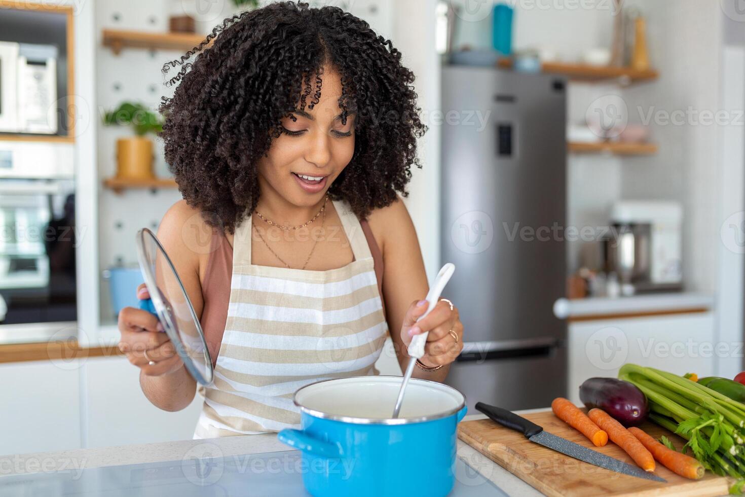 Photo of pretty African American woman holding cooking ladle spoon while cooking soup with fresh vegetables in kitchen at home