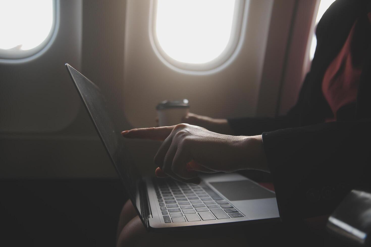 Blonde female tourist checking incoming notification on smartphone sitting on seat of airplane with netbook.Young businesswoman share media from telephone on laptop computer during plane flight photo