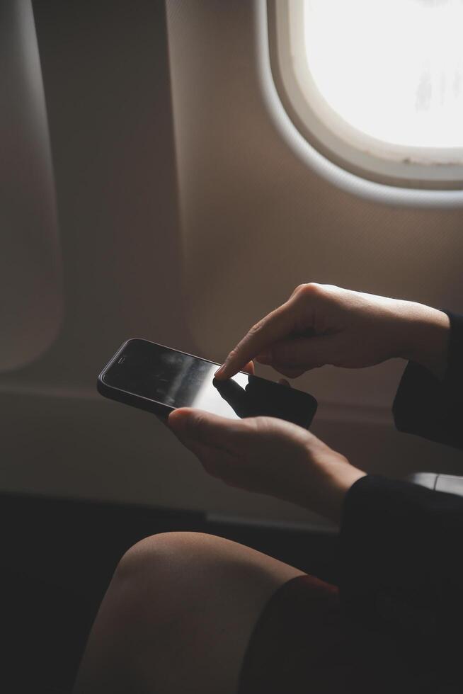Blonde female tourist checking incoming notification on smartphone sitting on seat of airplane with netbook.Young businesswoman share media from telephone on laptop computer during plane flight photo