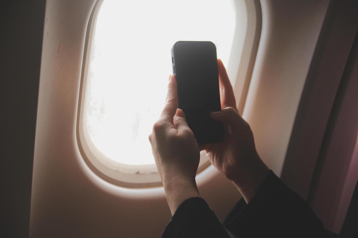 Blonde female tourist checking incoming notification on smartphone sitting on seat of airplane with netbook.Young businesswoman share media from telephone on laptop computer during plane flight photo