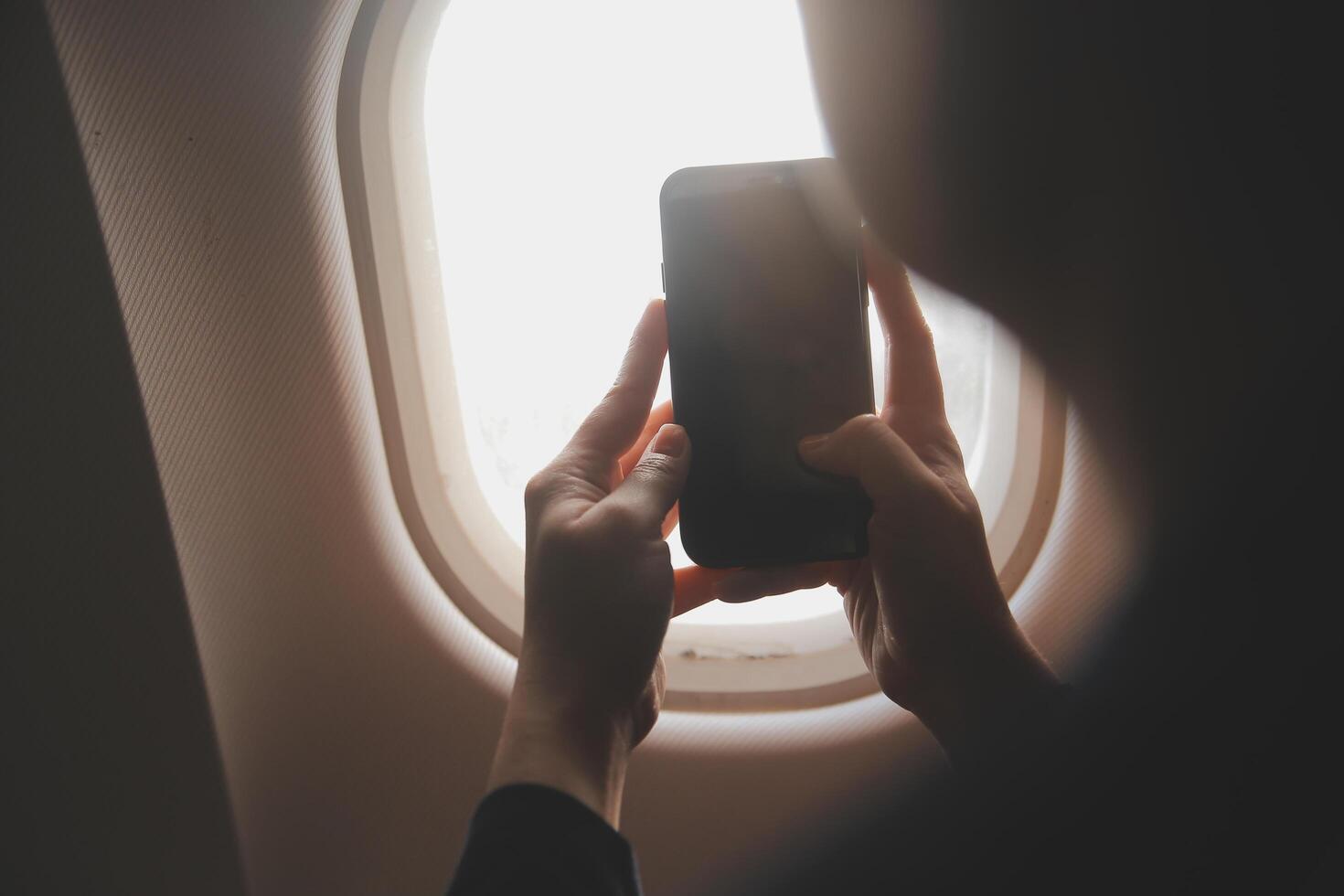 Blonde female tourist checking incoming notification on smartphone sitting on seat of airplane with netbook.Young businesswoman share media from telephone on laptop computer during plane flight photo